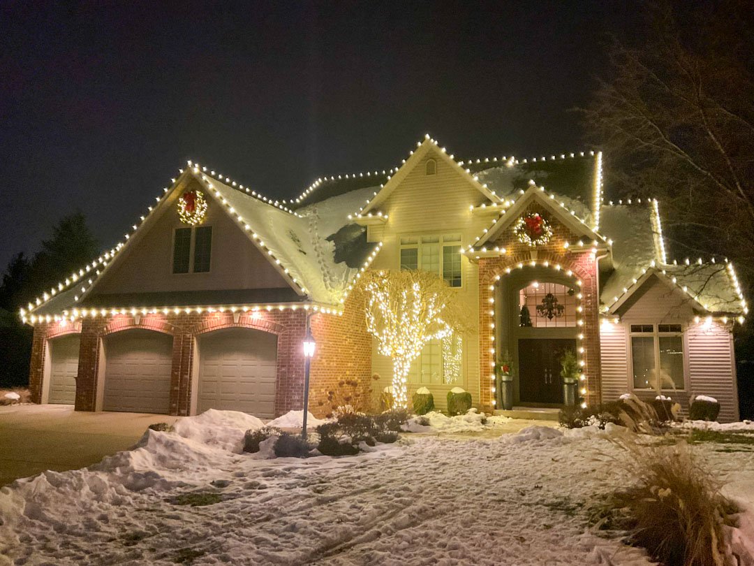 Warm white roofline lights, wreaths, and a light-wrapped tree on a snowy home