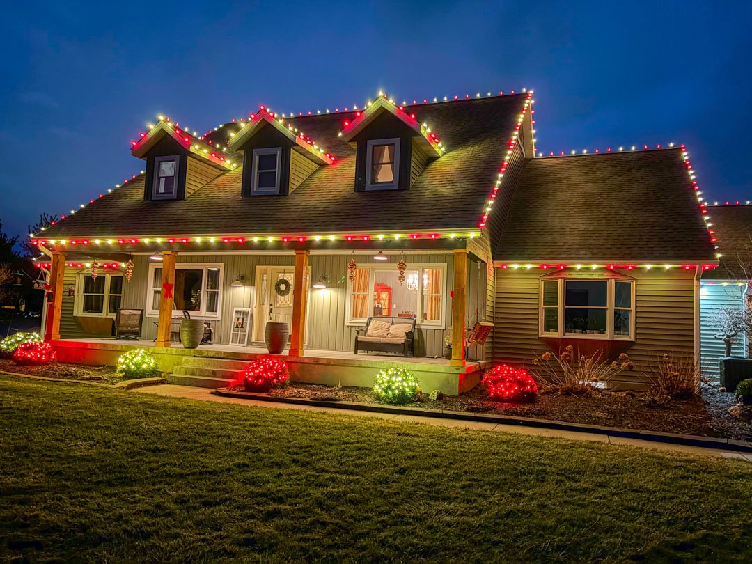 Colorful Christmas lights outlining a farmhouse roof, dormers, and porch